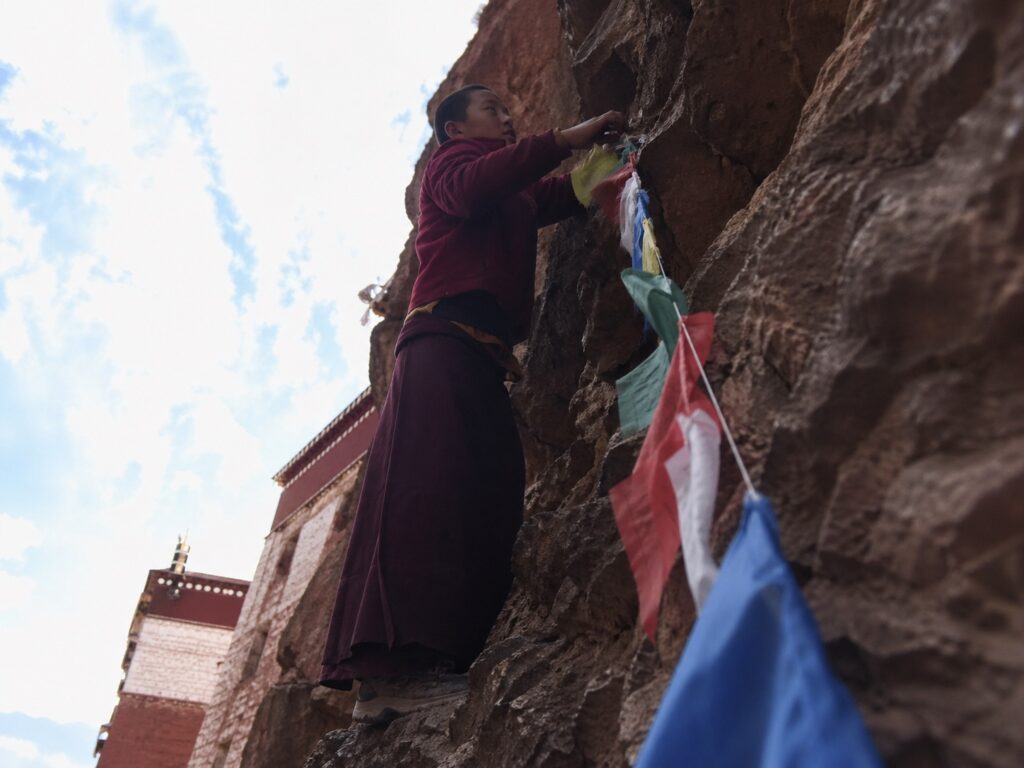 扎耶巴寺崖壁上的藏族男孩正在绑挂经幡，陡峭峭壁与彩色风马旗，西藏隐修圣地朝圣体验。